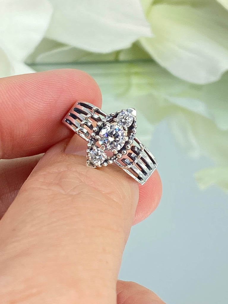 a person holding a ring with a flower in the background  a three stone ring with a White CZ center. Sterling Silver Art Deco Filigree, fine detail, Silver Embrace Jewelry, Xanadu D164