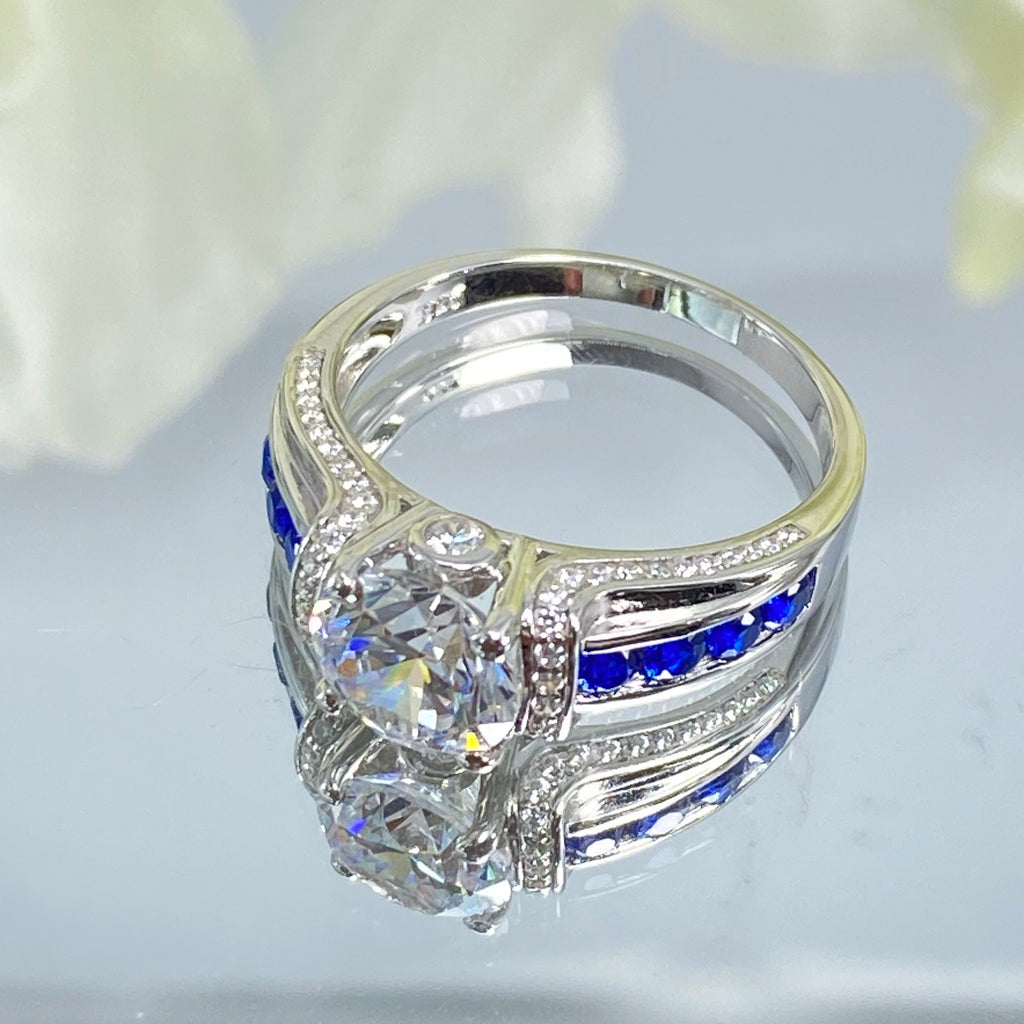 white CZ art deco ring, center stone is round cut white CZ, there are two trails of blue sapphire accents traveling up the sides of the band and two more trails of white CZs on each side of the band partially encircling the center stone, finally there are two accent white CZs on each of the remaining sides of the center stone, Sterling Silver Filigree, Silver Embrace Jewelry D220, Deco Inlay Ring