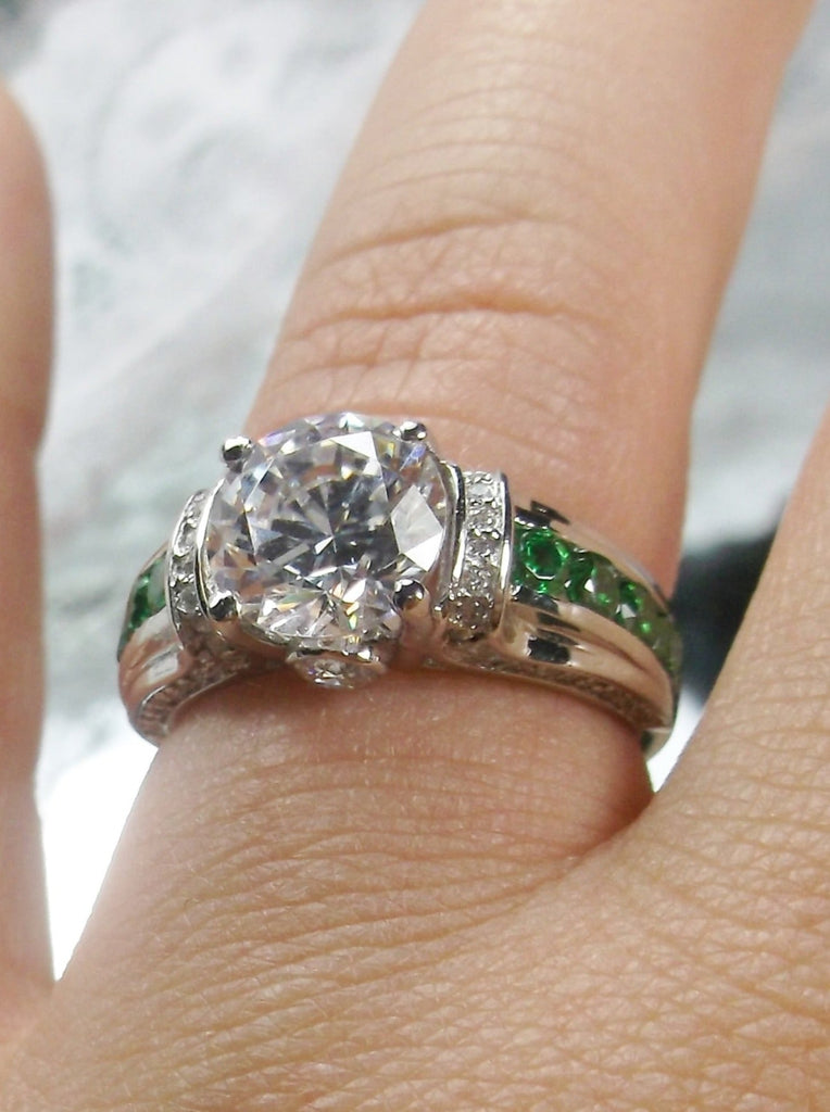 white CZ art deco ring, center stone is round cut white CZ, there are two trails of emerald green accents traveling up the sides of the band and two more trails of white CZs on each side of the band partially encircling the center stone, finally there are two accent white CZs on each of the remaining sides of the center stone