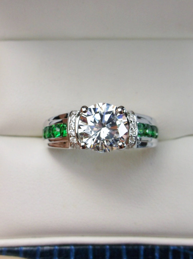 white CZ art deco ring, center stone is round cut white CZ, there are two trails of emerald green accents traveling up the sides of the band and two more trails of white CZs on each side of the band partially encircling the center stone, finally there are two accent white CZs on each of the remaining sides of the center stone