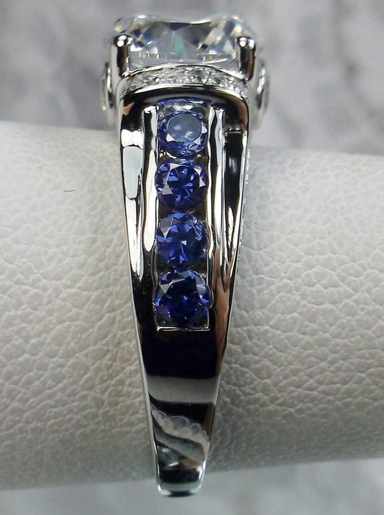 white CZ art deco ring, center stone is round cut white CZ, there are two trails of blue sapphire accents traveling up the sides of the band and two more trails of white CZs on each side of the band partially encircling the center stone, finally there are two accent white CZs on each of the remaining sides of the center stone