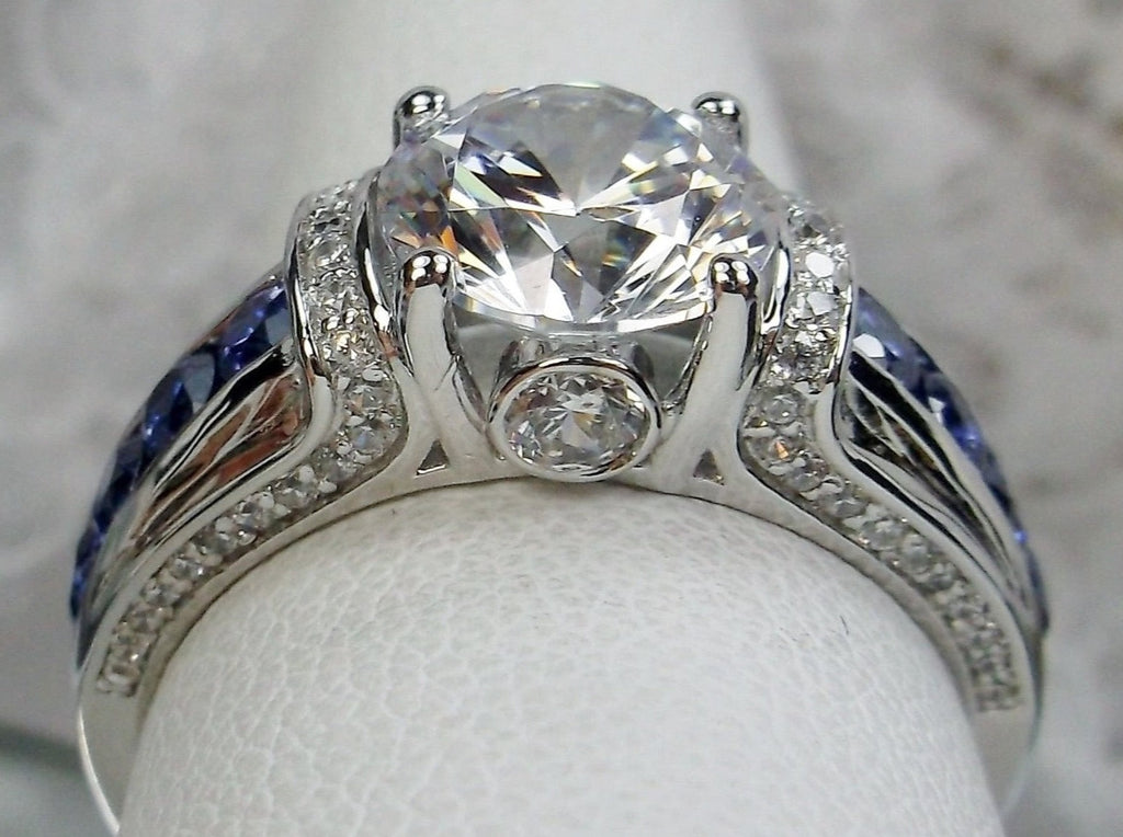 white CZ art deco ring, center stone is round cut white CZ, there are two trails of blue sapphire accents traveling up the sides of the band and two more trails of white CZs on each side of the band partially encircling the center stone, finally there are two accent white CZs on each of the remaining sides of the center stone