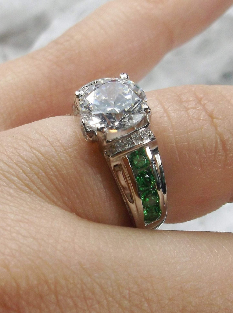 white CZ art deco ring, center stone is round cut white CZ, there are two trails of emerald green accents traveling up the sides of the band and two more trails of white CZs on each side of the band partially encircling the center stone, finally there are two accent white CZs on each of the remaining sides of the center stone
